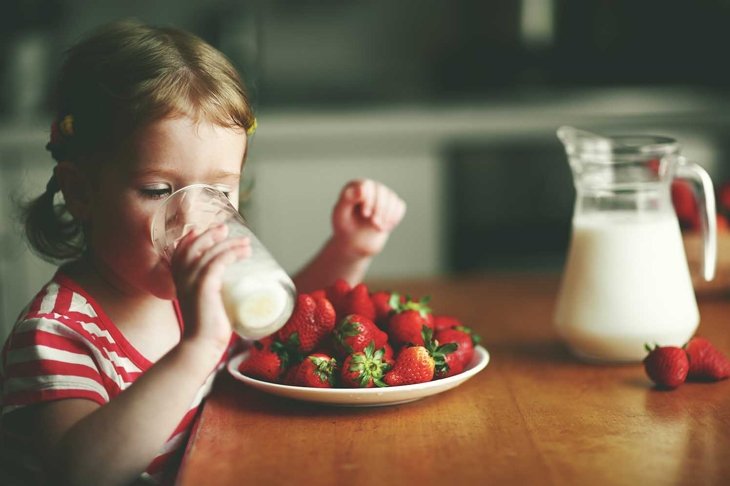 happy child girl drinks milk and eats strawberries in the summer home kitchen